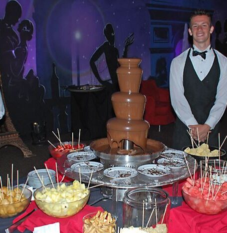 A smiling waiter stands beside a chocolate fountain surrounded by fruits and snacks on skewers at a decorated event.